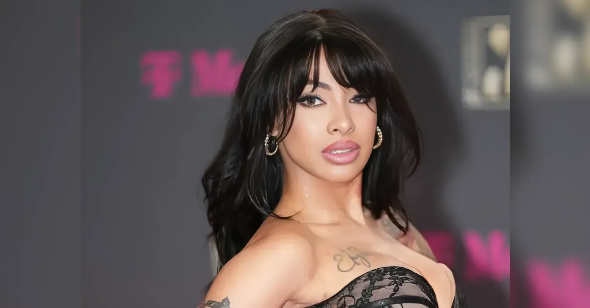 Portrait of a woman with long black hair and bold makeup wearing a black lace strapless top at a red carpet event, with gold hoop earrings.