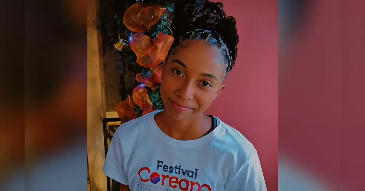 Smiling girl with a braided updo wearing a light blue festival shirt, standing beside a pink wall and colorful decor.
