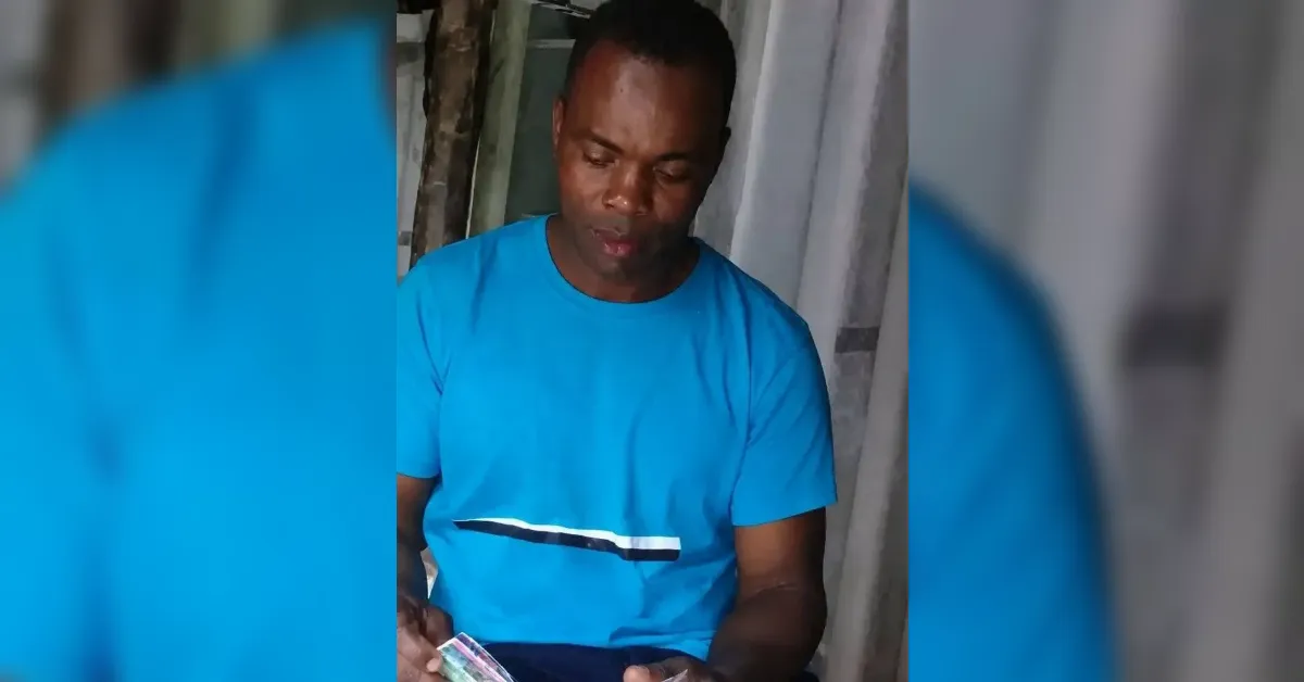 Man in a bright blue shirt sits indoors, counting money in his hands, looking down at the bills
