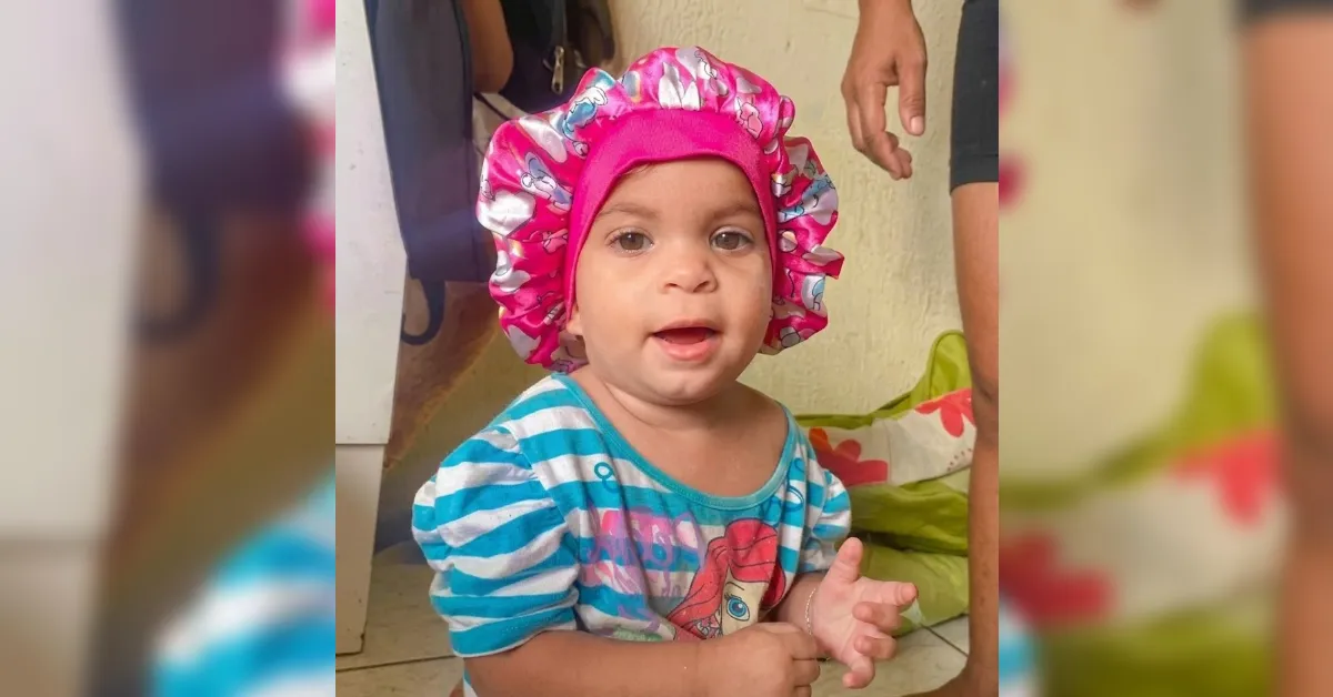 Baby girl wearing a bright pink frilly bonnet and a blue-striped shirt, indoors.
