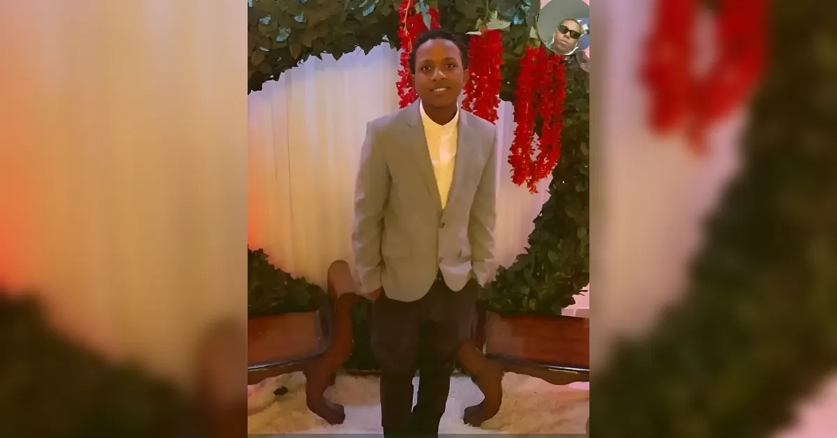 Young man in a light gray blazer and white shirt posing in front of a floral arch with red garlands.