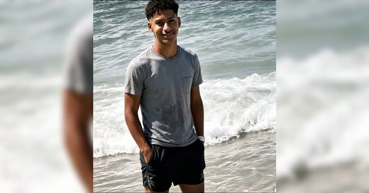 Man standing on the beach with waves behind him, wearing a gray t-shirt and black shorts, smiling on camera.