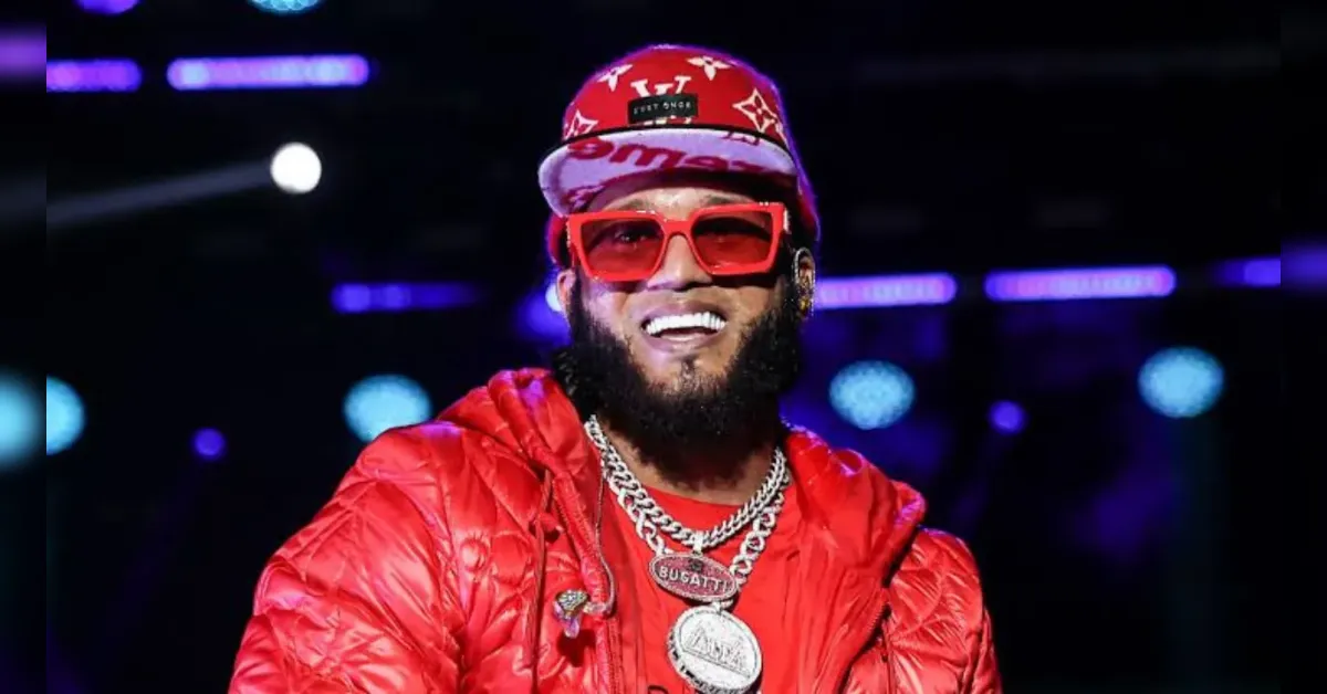 Smiling man in a red puffer jacket and red cap with oversized red sunglasses on a stage with colorful lights behind him.