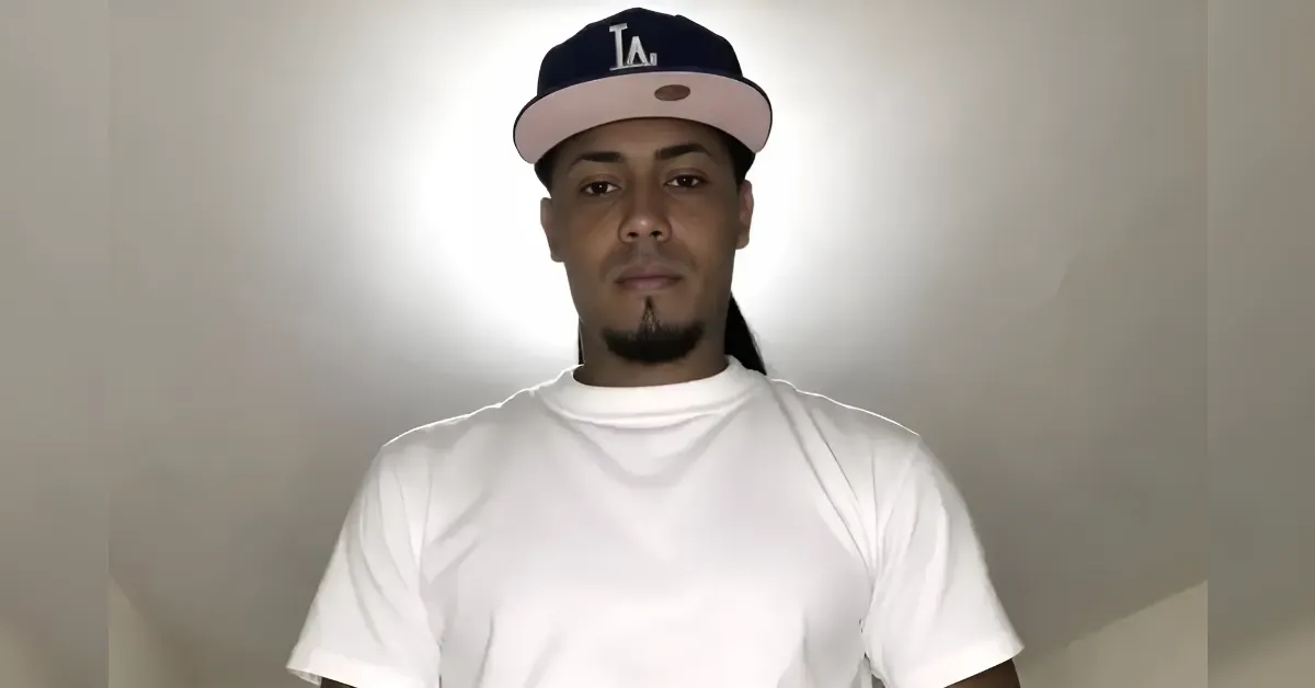Portrait of a man wearing a white T-shirt and a dark LA baseball cap, facing the camera.