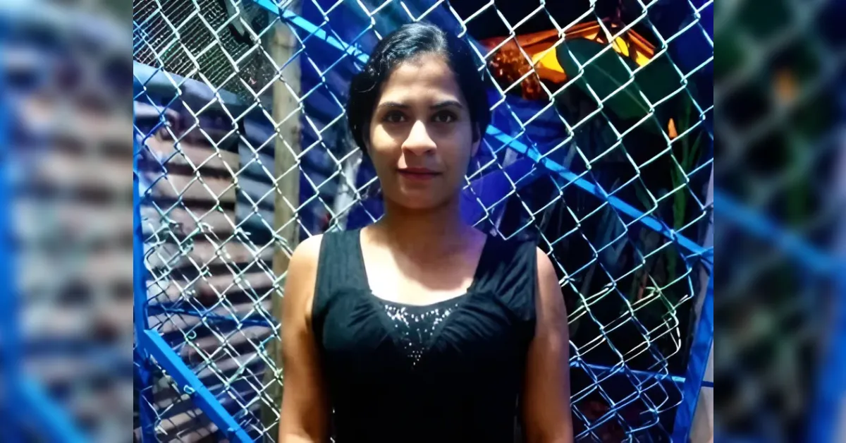 Woman standing in front of a chain-link fence, wearing a black sleeveless top, looking at the camera in a brightly lit setting.