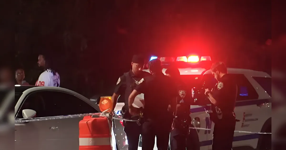 Nighttime scene with several police officers near a patrol car, red and blue lights flashing, and orange traffic barrels with police tape cordoning off the area.