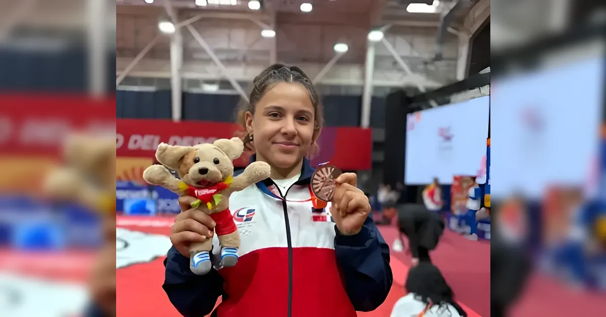 Young girl athlete in a red, white, and blue jacket holds a bronze medal and a stuffed dog in a sports arena.