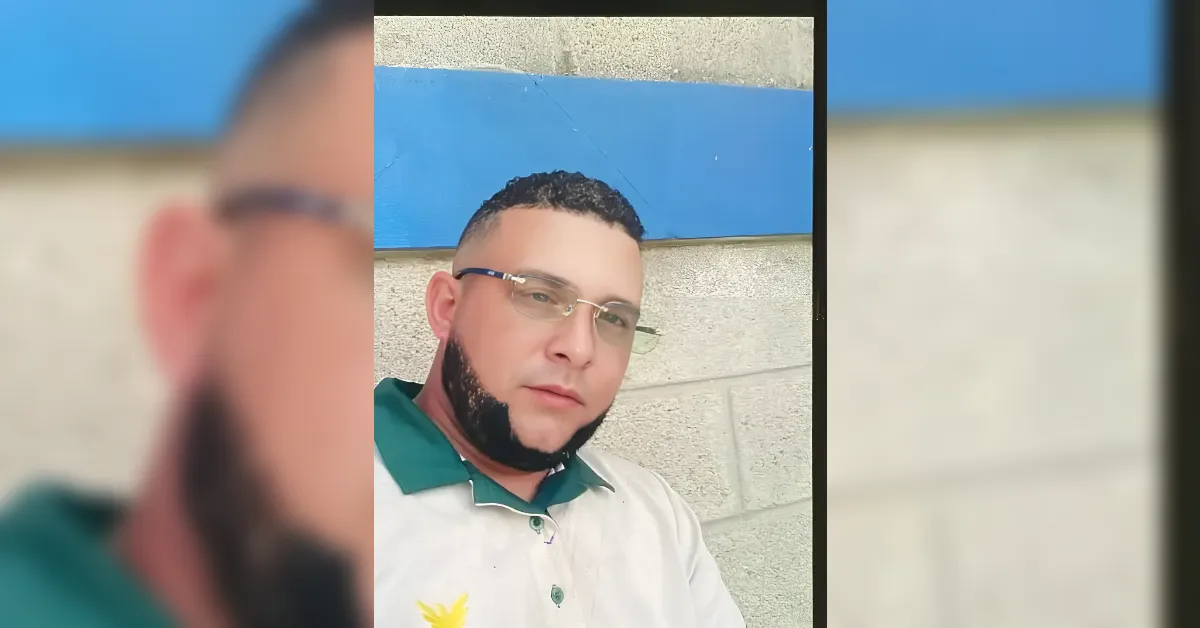 Portrait of a man with short dark hair, glasses, and a trimmed beard, wearing a green-collared polo, standing against a gray brick wall with a blue panel above.