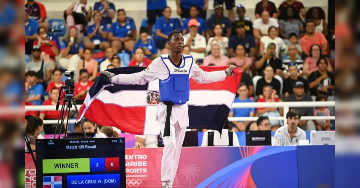 Taekwondo athlete in white uniform and blue chest guard stands on mat, arms outstretched holding the Dominican Republic flag as the winner with a crowd watching and a scoreboard showing 2-0.