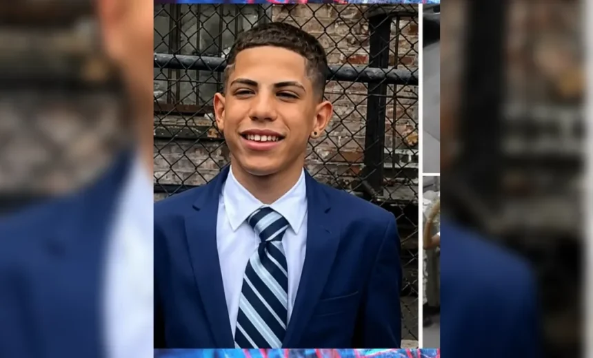 Young man in a navy suit and striped tie smiling in front of a chain-link fence.