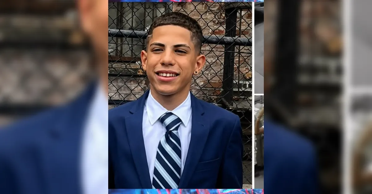 Young man in a navy suit and striped tie posing outdoors in front of a chain-link fence, smiling for the camera.
