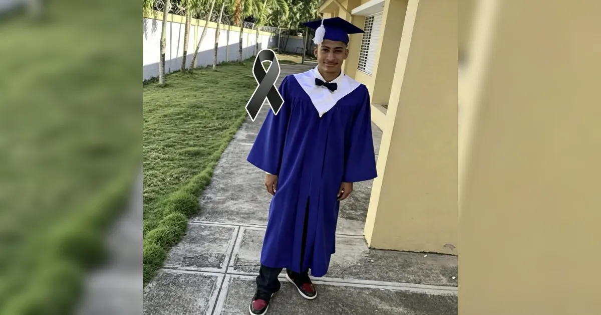 Young man in a blue graduation gown and cap posing on a sidewalk outside a yellow building.