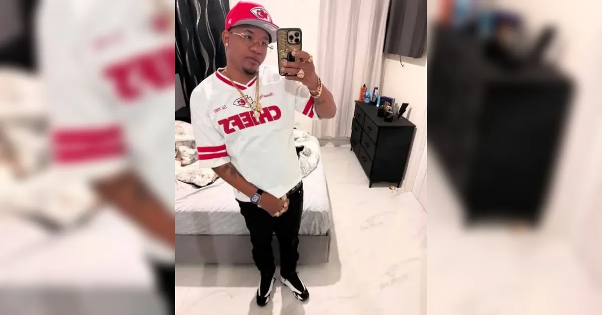 Man posing for a mirror selfie in a modern bedroom, wearing a white and red sports jersey, cap, and gold jewelry.
