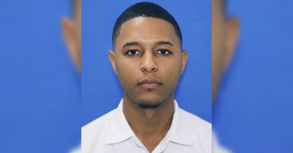 Portrait of a young man with short black hair and light stubble, wearing a white collared shirt against a blue background, with a neutral expression.