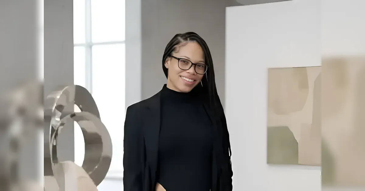Smiling professional woman with glasses in a black outfit stands in a bright art gallery near abstract sculptures and contemporary art.