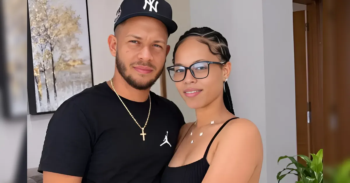 A couple taking a selfie indoors: man in a black t‑shirt and NY cap, woman with glasses and a black top beside him.
