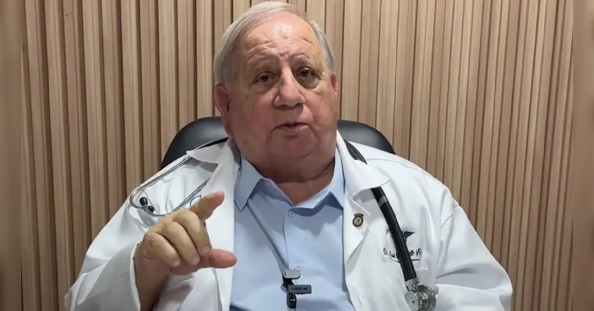 Older male doctor in a white coat with a stethoscope, pointing as he speaks in a medical office setting.