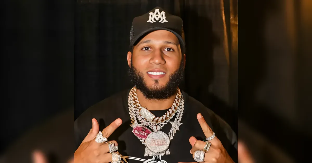 Portrait of a man in a black cap and multiple silver chains, wearing a heavy pendant and rings, flashing peace signs with both hands.