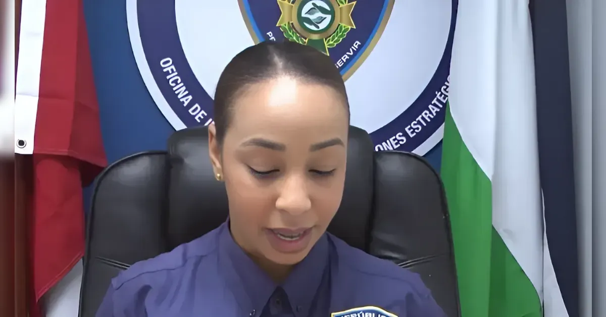 Female officer in a blue uniform seated at a desk, reading aloud with a police emblem and flags in the background.