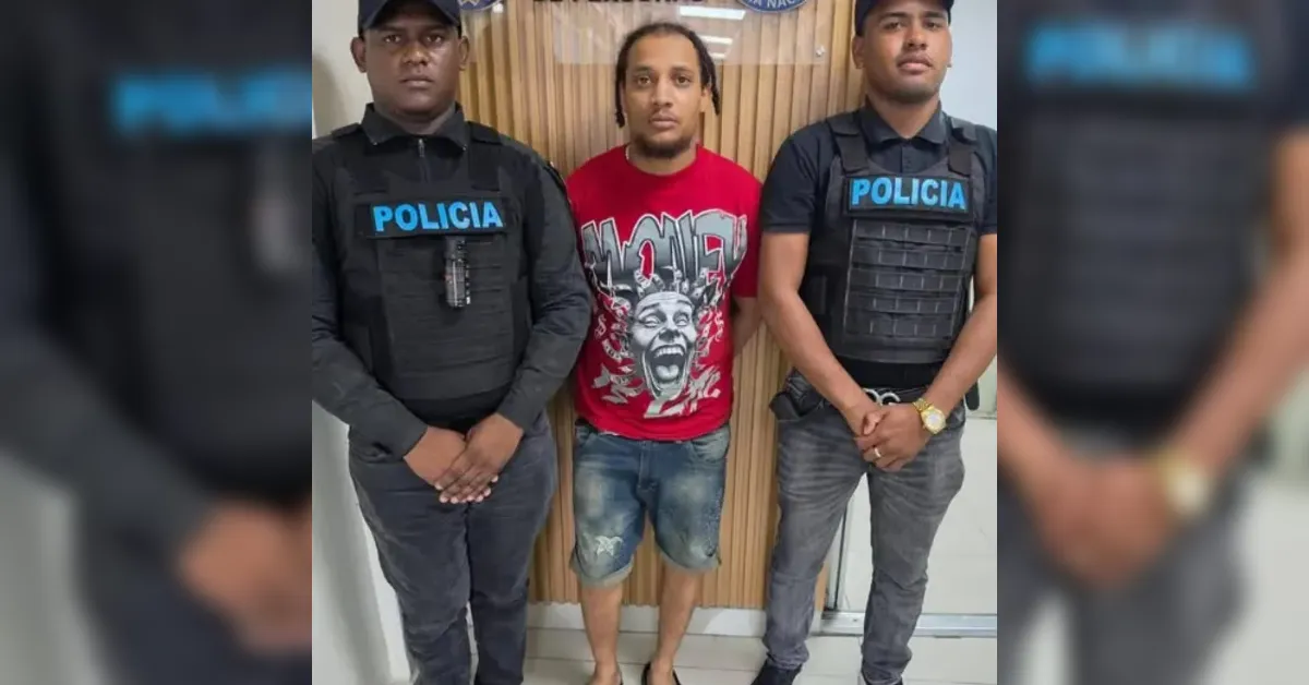 Man in a red graphic t-shirt stands between two police officers in bulletproof vests indoors.
