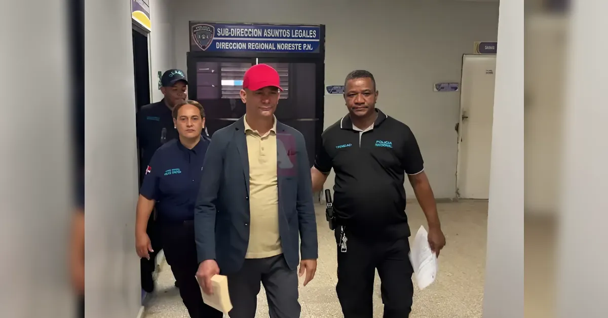 Man in a yellow polo and blue blazer with a red cap walking down a police hallway escorted by officers; sign reads legal affairs office in background.