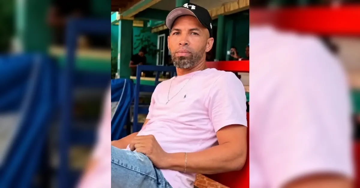 Man wearing a light pink t-shirt and cap sits on a red bench outdoors, looking at the camera with a neutral expression.