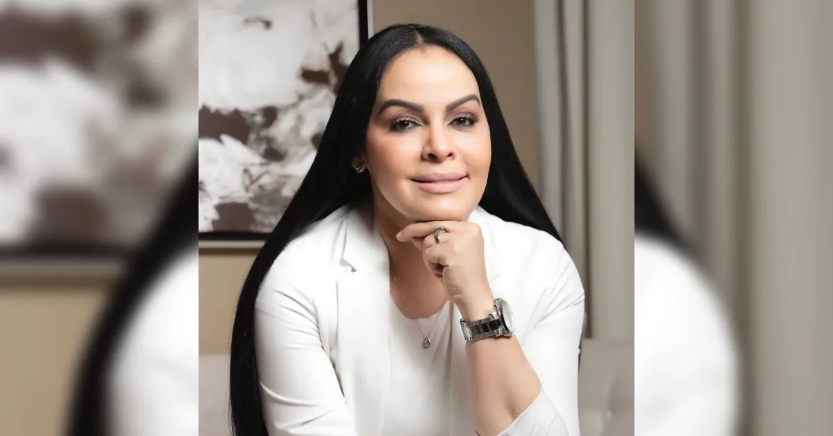 Portrait of a smiling woman with long black hair, wearing a white blazer and a watch, resting her chin on her hand.