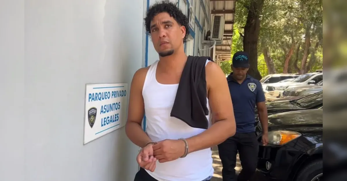 Man in a white sleeveless shirt with a towel over his shoulder stands with hands bound in front, escorted by a security officer outside a parking area.