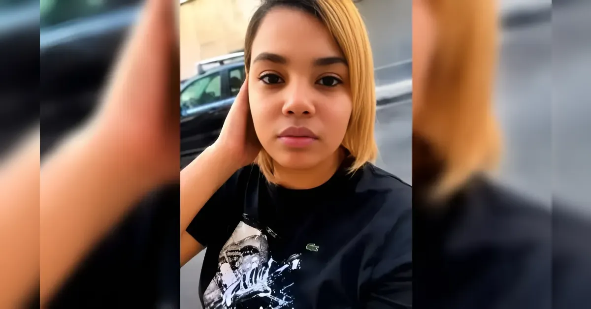 Woman with short blond hair wearing a black graphic t-shirt, looking at the camera with one hand resting on the side of her head, outdoors on a street.