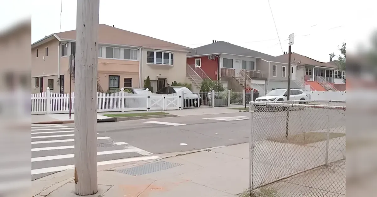 Suburban street scene with pastel houses, white fences, a crosswalk, and a metal utility pole in the foreground; a few cars parked along the curb.