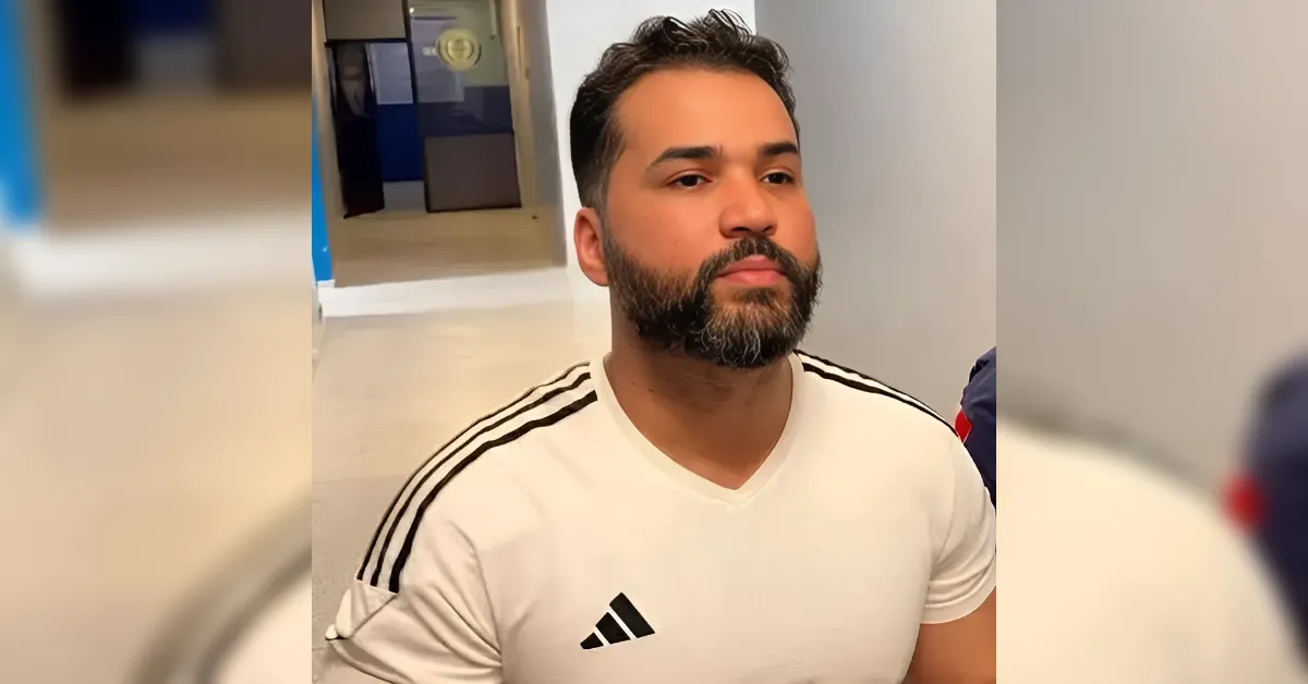Portrait of a man with dark hair and beard wearing a white Adidas t-shirt with black stripes, seated indoors and looking to the left