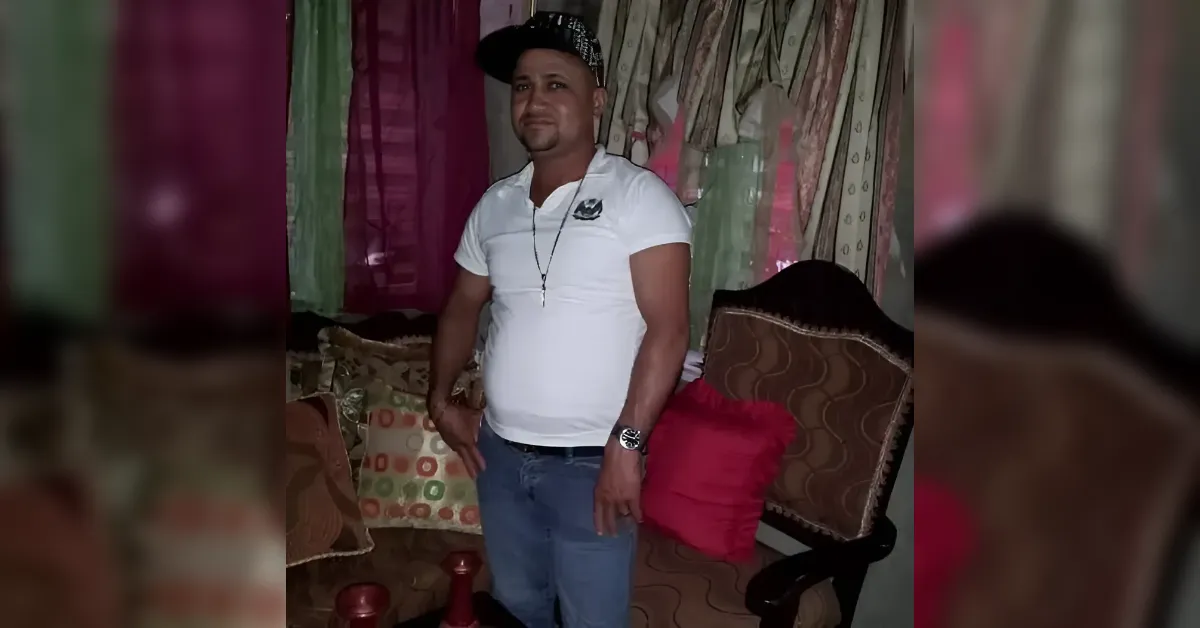 Man wearing a white polo, cap, necklace, and watch stands in a living room with patterned curtains and brown upholstered chairs.