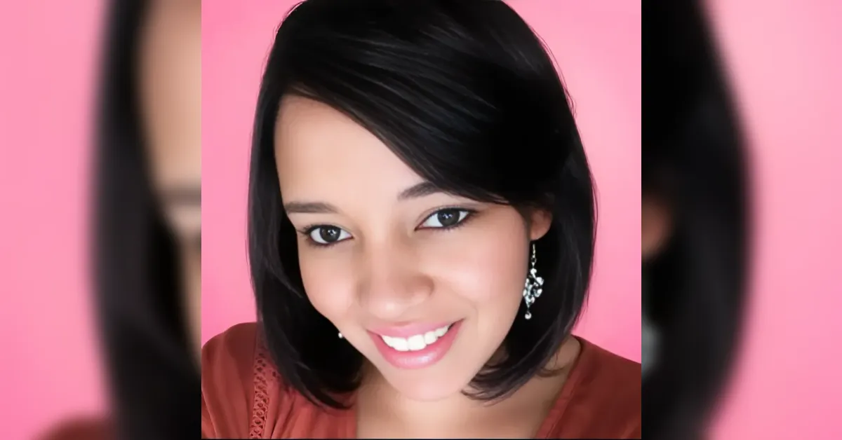 Smiling woman with short black hair against a pink background, wearing dangling earrings and a coral top.
