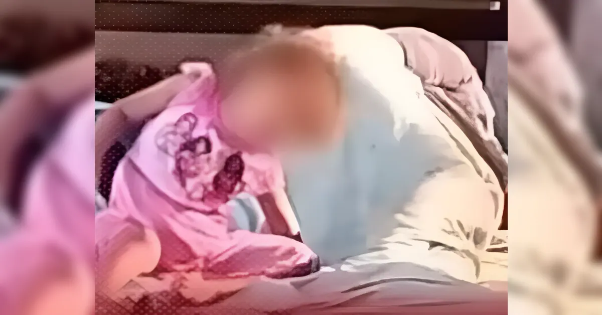 Baby in a pink outfit with a bib, lying on a bed with blankets nearby (blurry foreground). Central subject is a young child.