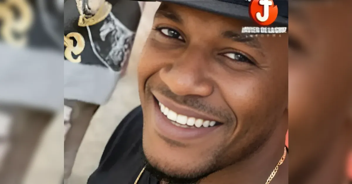 Close-up of a smiling man wearing a black shirt, a hat, and a gold chain; cap is tilted back.