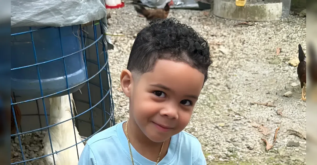 Boy with short curly hair in a light blue shirt smiles at the camera outdoors beside a blue metal chicken coop and poultry area.