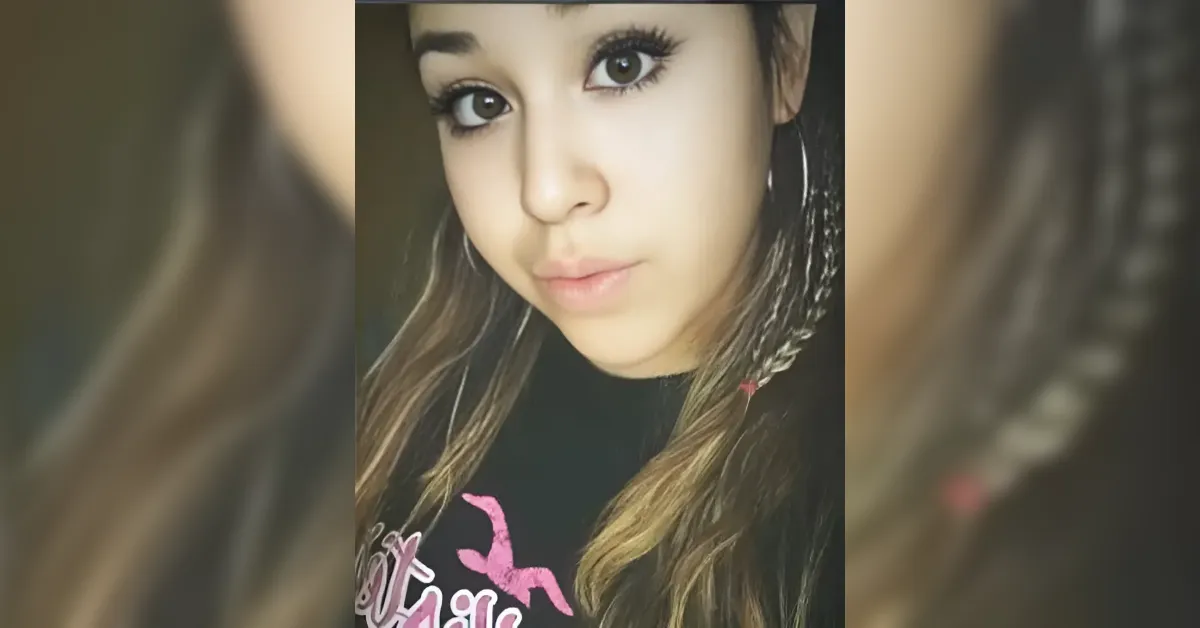 Close-up portrait of a young woman with long brown hair and a braid, wearing hoop earrings and a black shirt with pink lettering, looking at the camera.