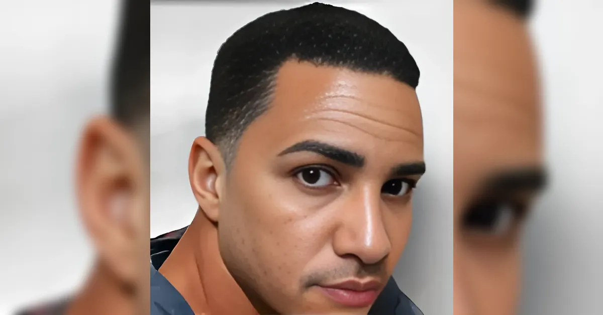 Close-up portrait of a man with short black hair looking at the camera, against a light background.
