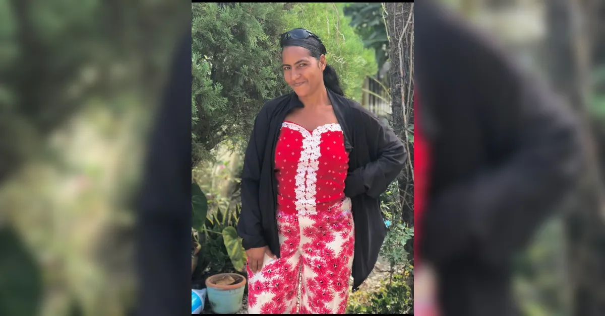 Woman standing outdoors in a red and white floral dress with a black jacket, hands on her hips, smiling at the camera, with greenery in the background.