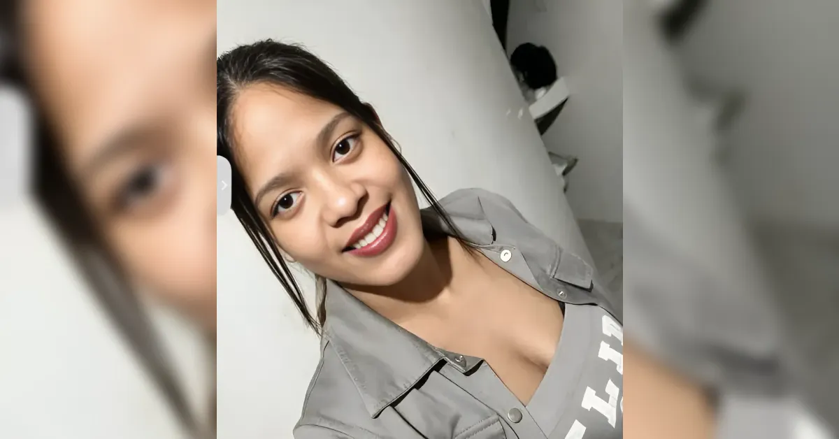 Smiling woman taking a selfie against a white wall, wearing a gray button-down shirt.