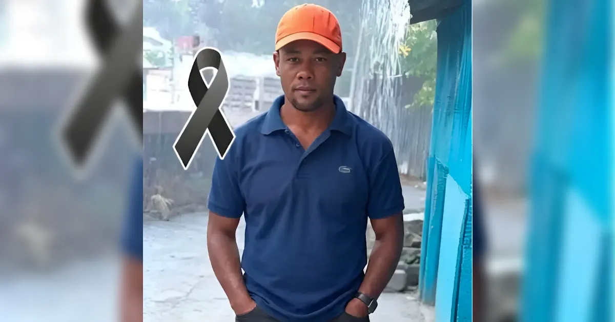Adult man in a blue polo and orange cap stands outdoors with hands in pockets; a black awareness ribbon graphic overlays the left chest area, suggesting a tribute or awareness message.