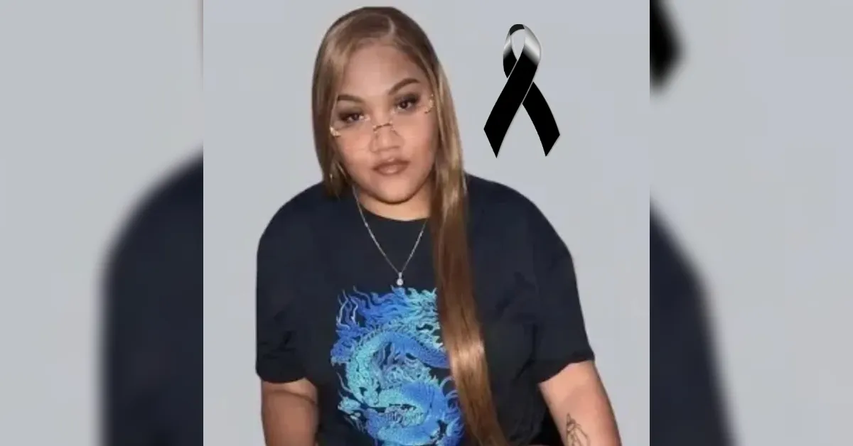 Woman with long light brown hair wearing a black T‑shirt with a blue dragon design; a black mourning ribbon appears in the top right corner.