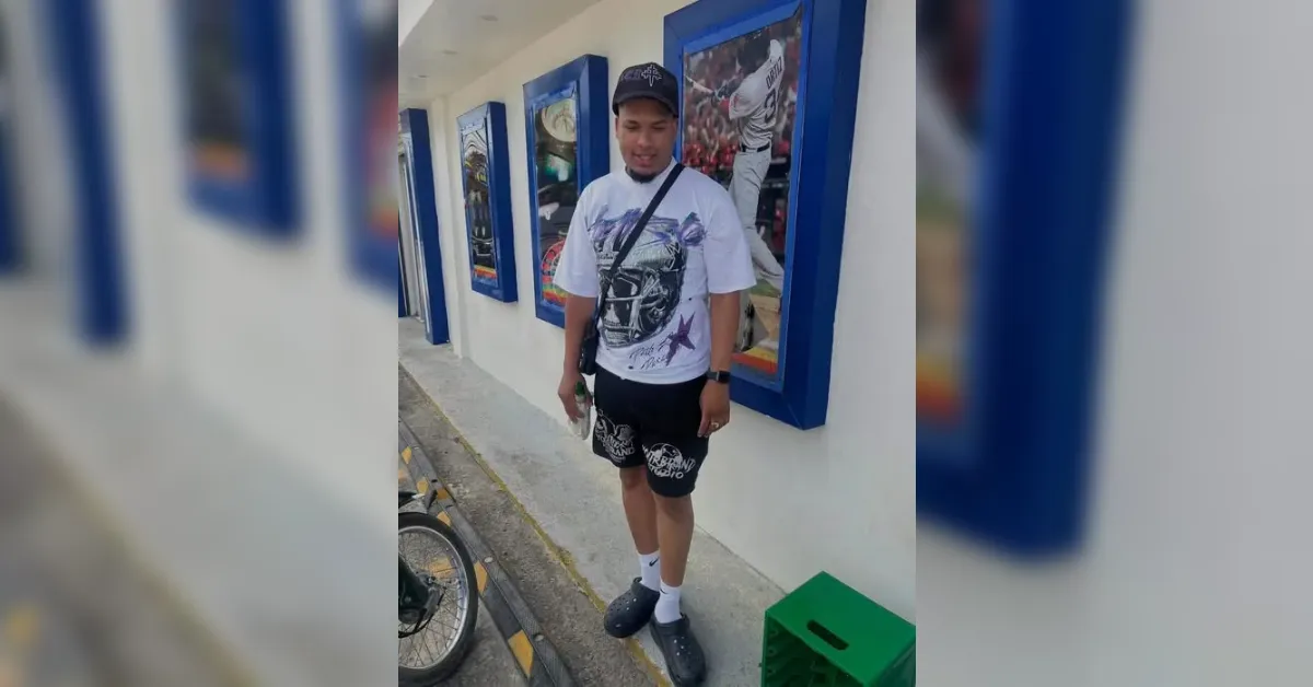 Man wearing a white graphic T-shirt, black shorts, cap, and crossbody bag stands on a sidewalk beside blue-framed posters on a white wall, holding a drink.