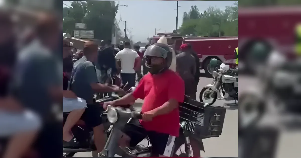 Man in a red shirt wearing a helmet rides a scooter with a basket, amid a crowd on a street with emergency vehicles in the background.