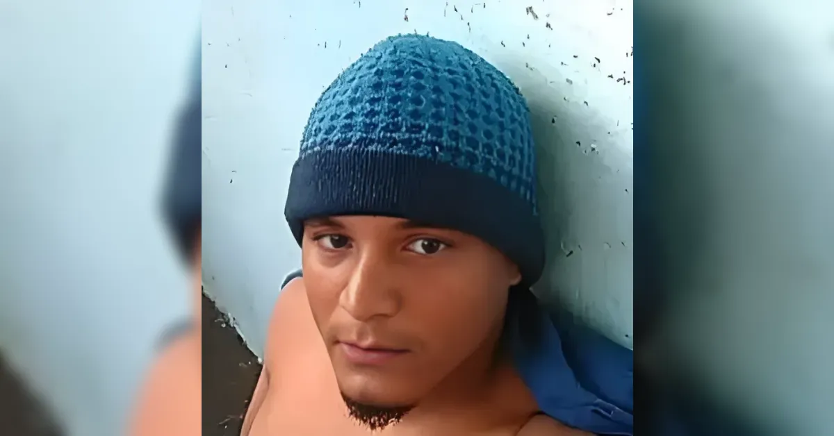 Young man wearing a teal knit beanie and blue scarf leaning against a light blue wall, looking at the camera.