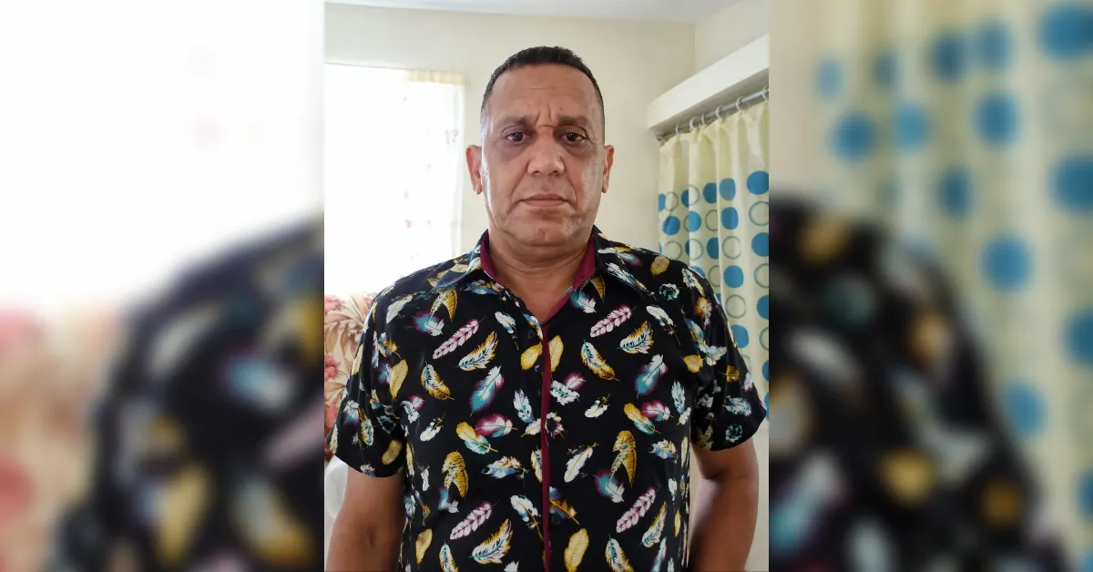 Man indoors facing the camera, wearing a dark shirt with colorful feathers, with a patterned shower curtain behind him in a bright room.