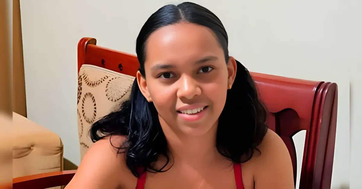 Young woman with dark hair tied back, smiling at the camera while sitting on a wooden chair indoors.