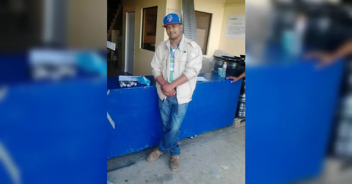Man wearing a blue cap and beige jacket stands with hands clasped in front of a blue counter inside a building, posing for the photo.