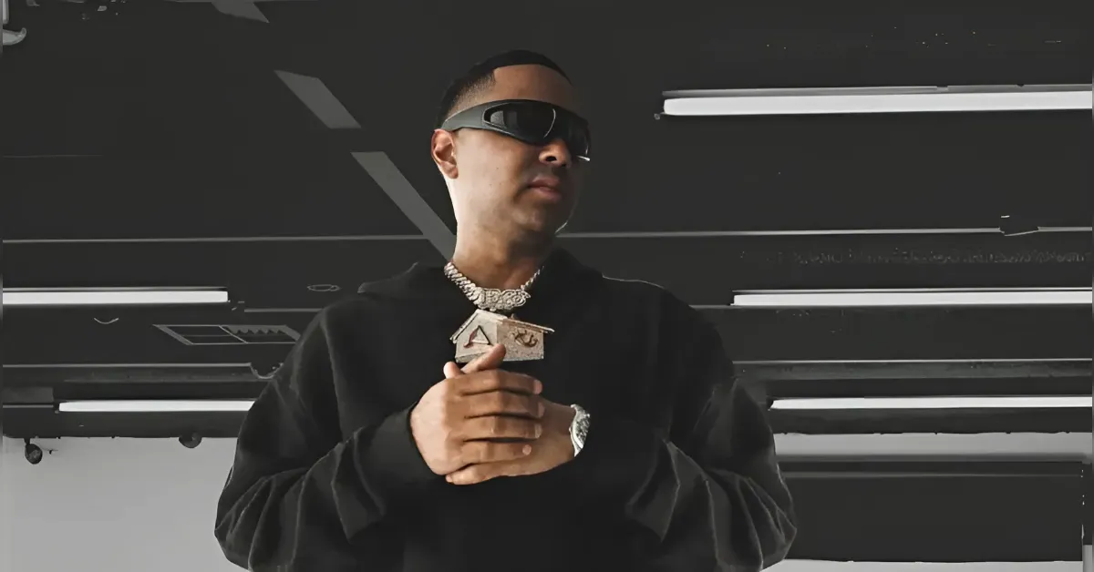 Man in a black hoodie and sunglasses stands in a dim studio, hands clasped, wearing a heavy diamond necklace and pendant.