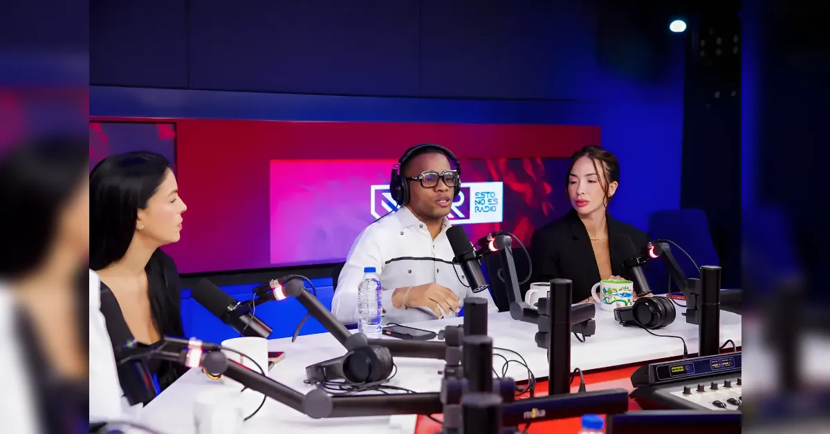 Three hosts sit at a radio studio desk with microphones, headphones, and a mixer under blue and red lighting.